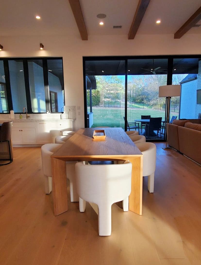 Modern dining area featuring a wooden table and white chairs, large windows with outdoor view.