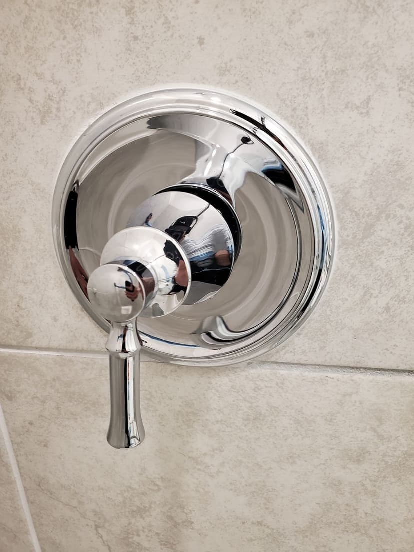 Chrome shower valve on textured tile wall, reflecting light and surroundings.